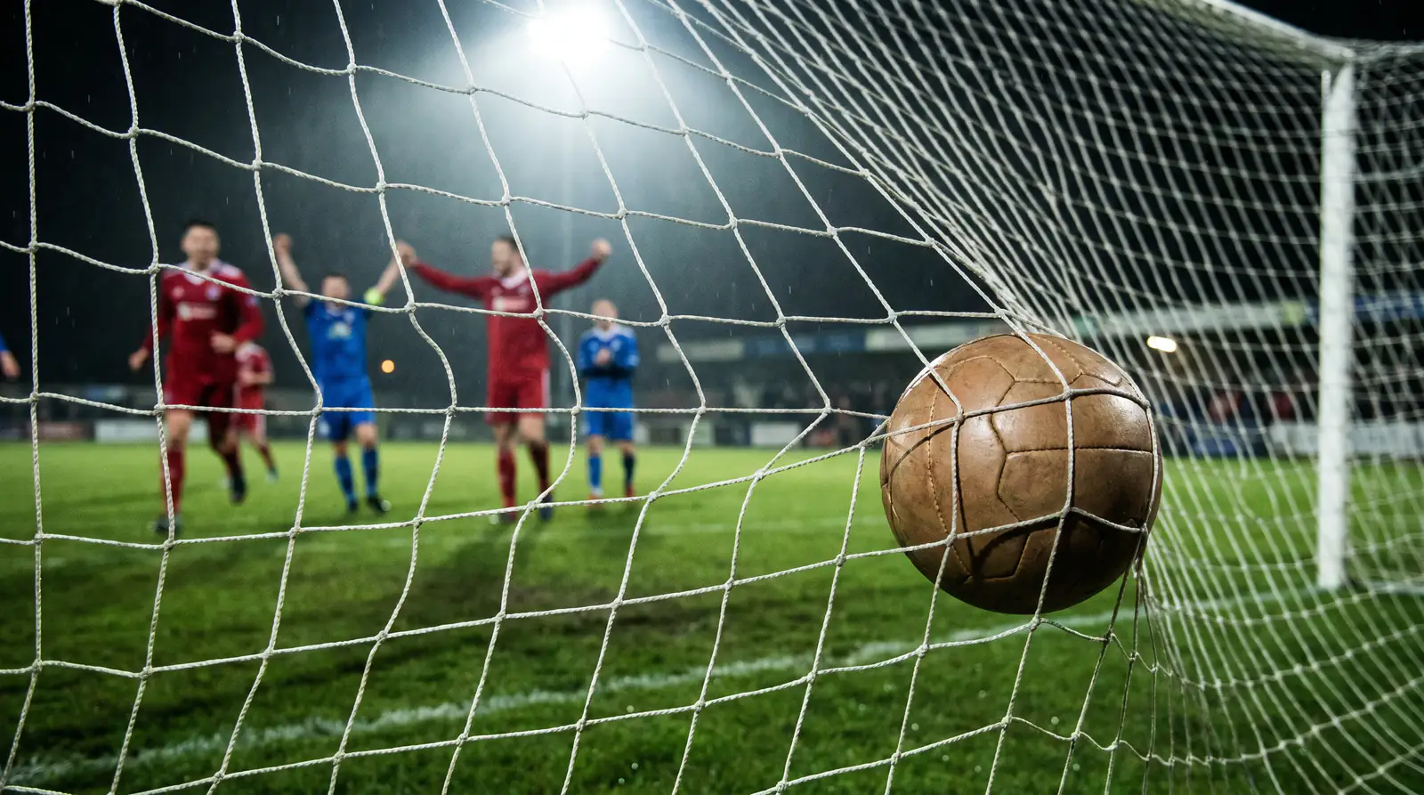 Football hitting the back of the net during a goal in a floodlit stadium